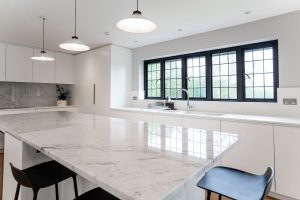 Modern well-lit kitchen featuring black Crittall Homelight Windows along one wall. The kitchen is white marble and features several hanging lights. 
