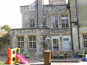  A slightly off-center, medium-distance shot of the back of a large stone house on a sunny day. A patio extends from the house with a wooden table and chairs set. A large brown plastic wheeled garbage can stands near the center of the patio. In the bottom left corner, a brightly colored plastic children's slide and playset is visible.