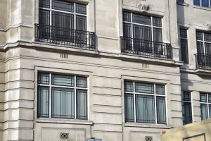 A wide, eye-level shot of a building facade with three rows of windows. The building is made of light-colored stone, possibly from the 1930s or 40s. The windows have multi-paned steel frames, with the upper panes having a more decorative, geometric grid pattern. The middle row of windows has balconies with intricate steel railings. The building has a decorative Art Deco motif of a shell or fan carved into the stone between the two upper-level windows.